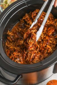 mengeluarkan daging babi dari crockpot untuk membuat Sandwich Babi Tarik Slow Cooker